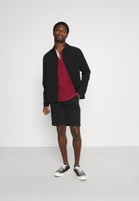 Svart jakke, rød polo med hvite detaljer, svarte shorts og svarte joggesko. Modellen står med en hånd på hoften mot en ensfarget bakgrunn.