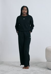 Črni komplet oblačil za sproščanje s puloverjem z dolgimi rokavi in ujemajočimi hlačami. Pulover ima okrogel izrez; hlače z elastiko v pasu in okraski iz cofkov.