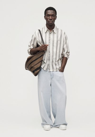 Un homme porte une chemise rayée boutonnée, un jean clair ample et des baskets blanches. Il porte un grand sac bandoulière marron à rayures, sur un fond uni.