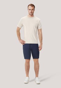 Beige bomulls t-shirt, marinblå shorts och vita sneakers. Modellen står med en hand på höften och visar upp en avslappnad sommaroutfit.