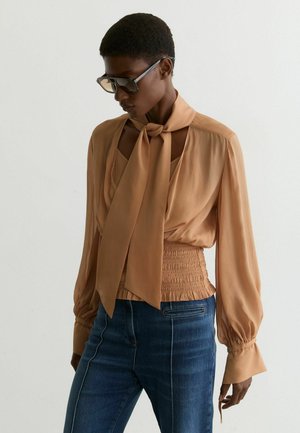 Persoană cu păr scurt, purtând ochelari de soare mari, o bluză bronzată cu mâneci lungi și cravată la gât, și blugi albaștri cu talie înaltă, stând în fața unui perete simplu.