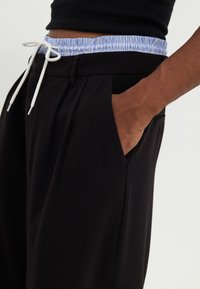 Pantalons noirs avec un design plissé, comprenant une ceinture élastique rayée bleu clair et un cordon blanc. Inclut des poches latérales.