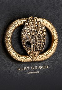 Czarna skórzana torebka na ramię z złotym zapięciem w kształcie dłoni, z misternymi detalami, otoczona okrągłym, gwiazdkowym wzorem oraz tłoczonym logo.