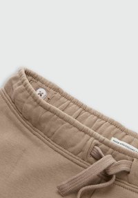 Beige bomuldsbukser med snøre i taljen, der har en ribbet elastik, med en knaplukning og et stofmærke, der siger "lavet med økologisk". Glat tekstur.