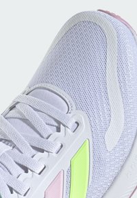 Chaussure de sport blanche avec une tige en maille, des accents texturés en rose et vert, et un design de semelle blanche incurvée. Les lacets sont visibles.