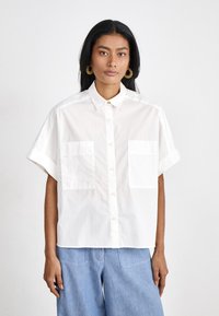 White short-sleeve button-up shirt made of cotton, featuring a collared neckline and two front pockets, worn over blue denim trousers.