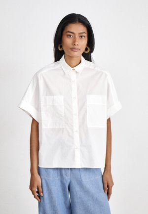 White short-sleeve button-up shirt made of cotton, featuring a collared neckline and two front pockets, worn over blue denim trousers.