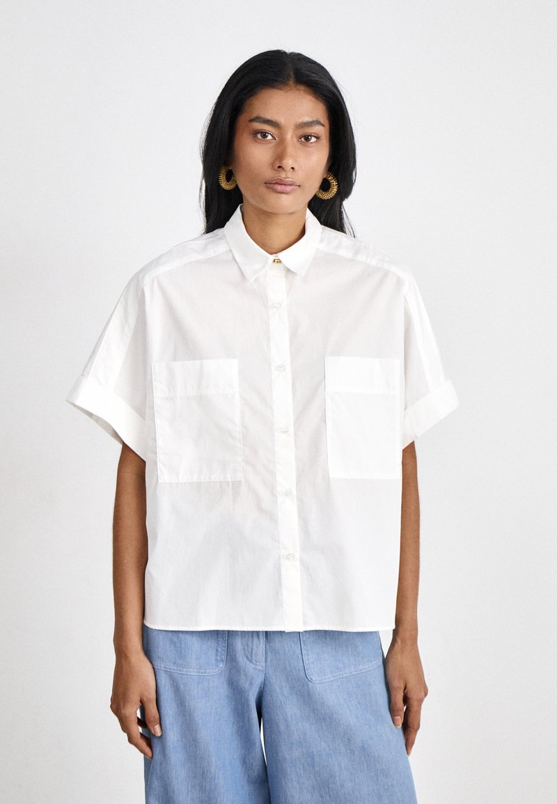 White short-sleeve button-up shirt made of cotton, featuring a collared neckline and two front pockets, worn over blue denim trousers.