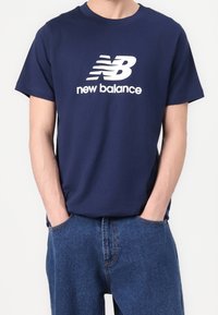 Tmavě modré bavlněné tričko s krátkým rukávem, na kterém se nachází velké bílé logo "NB" a text "new balance" umístěný přímo pod ním.