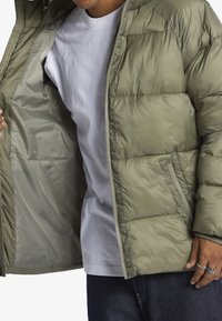 Grön puffjacka med dragkedja framtill, med en slät och blank yta, horisontella sömmar och sidofickor. Innerfodret är ljusare.