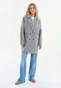 Žena stojí oblečená v nadrozmernom sivom kabáte s rybacími kosťami, svetlomodrých širokých džínsach a béžových nazúvacích topánkach, na bielom pozadí.