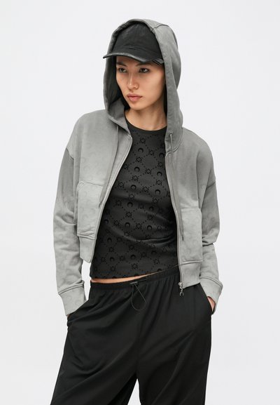 Γκρι κοντό φούτερ με κουκούλα, μπροστινή τσέπη και διακριτή υφή με σχέδιο. Φορεμένο πάνω από μαύρο εφαρμοστό τοπ με κυκλικά σχέδια και συνδυασμένο με φαρδιά μαύρη παντελόνια.