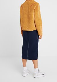 Person som bär en tanfärgad fluffig jacka, marinblå midikjol med vit söm, vita strumpor och vita sneakers, stående mot en enkel bakgrund.