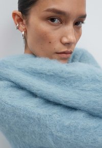 Giovane con capelli corti e scuri, indossa orecchini a cerchio d'argento e un morbido maglione azzurro chiaro e peloso, guarda di lato con un'espressione neutra.