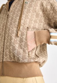 Brouwne rits-hoodie met een geometrisch patroon, ribgebreide manchetten en rand. De stof heeft een zachte textuur, met een logoborduring op de borst.