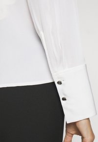 Bílá halenka s hladkou texturou na rukávech, se zdařilým manžetovým zakončením a třemi černými dekoračními knoflíky, s jemným leskem.