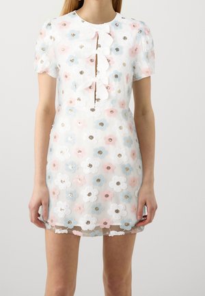 Femme portant une mini-robe blanche à manches bouffantes, ornée de motifs floraux pastel rose, bleu et blanc avec trois nœuds blancs sur le devant.