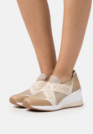 Piedi con indosso sneaker beige slip-on con suole in gomma bianca e fasce elastiche incrociate con il marchio "KORS".