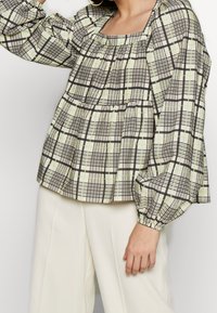 Blouse à carreaux vert clair et noir avec un décolleté carré, manches longues bouffantes et détails froncés à la taille.