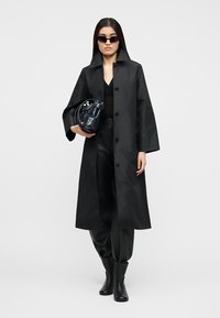 Γυναίκα με μακρύ μαύρο παλτό, παντελόνια, μπότες, γυαλιά ηλίου, κρατώντας σκοτεινή τσάντα, στέκεται μπροστά σε απλό ανοιχτό φόντο.