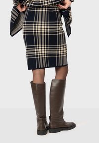 Spódnica w kratę do kolan w odcieniu granatu z beżowymi liniami, noszona z brązowymi skórzanymi botkami do kolan z niskim obcasem w kształcie bloku i teksturalnymi detalami.
