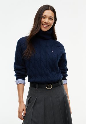 Jeune femme souriante portant un pull à col roulé bleu marine par-dessus une chemise rayée et une jupe grise plissée avec une ceinture noire sur fond blanc.