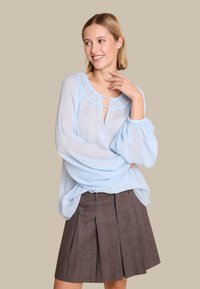 Lyseblå, gjennomsiktig bluse med volanger i halsen og nøkkelhullsdetalj, kombinert med en brun rutete plissert skjørt. Myk tekstur og løs passform.