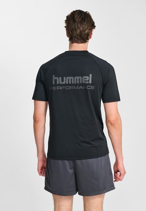 Man met de rug naar de camera, draagt een zwart "hummel performance" T-shirt en grijze sportshorts tegen een effen lichte achtergrond.