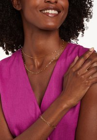 Goudlaag ketting en armband, gekoppeld ontwerp, gedragen met een roze mouwloze top. Gladde textuur, eigentijdse stijl, minimale accenten.