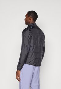 Man facing sideways wearing a black quilted jacket and light purple pants against a plain white background.