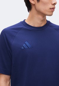 Άντρας που φοράει ένα απλό σκούρο μπλε T-shirt με ραμμένο σκούρο μπλε λογότυπο Adidas στο αριστερό στήθος, κοιτάζει προς τα δεξιά.