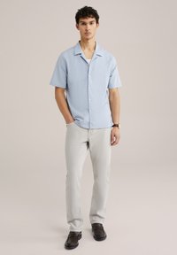 Camicetta azzurra a maniche corte con colletto, dotata di bottoni lungo la parte anteriore. Abbinata a pantaloni grigio chiaro e scarpe scure.