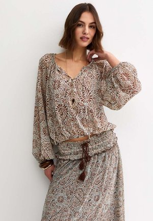 Kobieta ubrana w wzorzystą bluzkę z długim rękawem o luźnych zakończeniach oraz dopasowaną spódnicę, uzupełnioną warstwowymi naszyjnikami i bransoletkami.