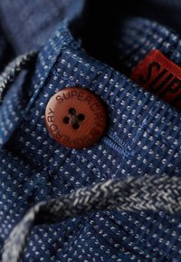 Tejido texturizado azul marino con costuras blancas, que presenta un botón redondo marrón con "SUPERDRY" grabado y un lazo tejido para el cierre.