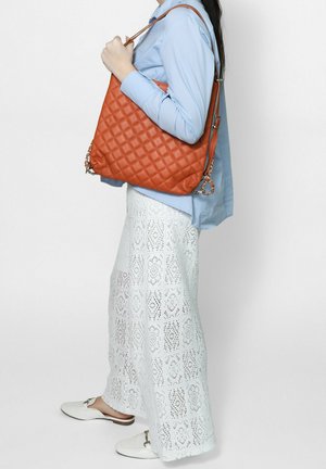 Donna con camicia blu e gonna di pizzo bianca che tiene una grande borsa trapuntata marrone sulla spalla, su uno sfondo semplice.