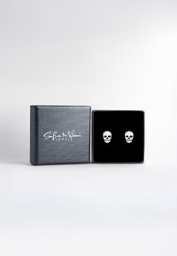 Schmuckkästchen mit schwarzem Samt-Futter und silbernen Totenkopf-Ohrhängern. Die Box hat eine strukturierte matte Oberfläche und ein weißes Logo.