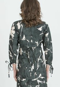 Langärmliges Kleid aus dunkelgrünem Stoff mit cremefarbenem Blumenmuster, in der Taille gebunden, mit gerafften Ärmeln und einer glatten Textur.