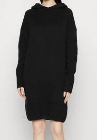 Abito a maglione nero con cappuccio, realizzato in materiale elasticizzato, presenta dettagli a costine sui polsini e sull'orlo, con scollatura a V e vestibilità comoda.