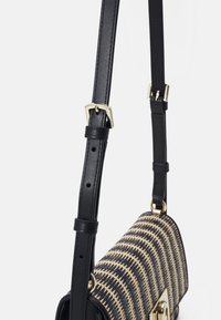 Bandoulière ajustable en cuir noir avec boucle dorée attachée à un sac bandoulière tissé noir et beige à motif.