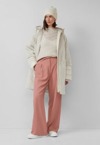 Oversized crèmekleurige jas over een gestructureerde gebreide top, gecombineerd met roze wijde broekspijpen en witte sneakers, geplaatst tegen een neutrale achtergrond.