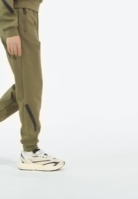Olivové nohavice s čiernymi akcentmi, so športovým pásom a bočnými vreckami, skombinované s bielymi a čiernymi teniskami.