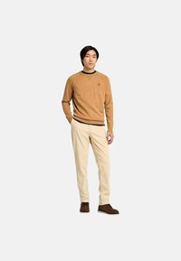 Maglione lavorato a maglia color beige con scollatura rotonda e piccolo logo, abbinato a pantaloni color crema e scarpe marroni.