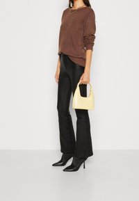 Brun stickad tröja, svarta utsvängda byxor, gul handväska med böjt handtag och svarta högklackade skor, visade mot en enfärgad bakgrund.