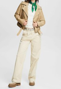 Skórzana motocyklowa kurtka w kolorze brązowym, biała sweter, beżowe szerokie spodnie i beżowe trampki. Zielony szalik zawiązany na szyi dodaje koloru.