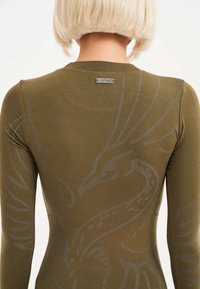 Oliwkowa bluzka z długim rękawem o gładkiej teksturze. Posiada subtelny szary wzór ptaka i małą metalową metkę przy dekolcie.