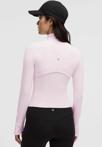 Jasnoróżowa, długosleeve'owa koszulka sportowa o dopasowanym kroju, z wysokim kołnierzem i przeszyciami z tyłu. Na głowie modela czarna czapka.