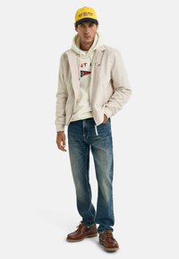Lichtbeige zip-up jas over een witte hoodie, blauwe jeans en bruin veter schoenen. De pet is geel met blauwe accenten en logo.