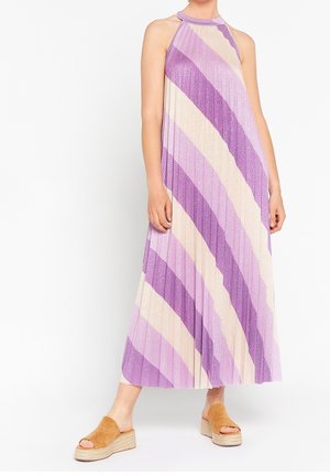 Kobieta ubrana w bezrękawnikową, plisowaną sukienkę maxi z przekątnymi paskami w kolorze lawendowym i beżowym, w połączeniu z jasnobrązowymi zamszowymi sandałami na platformie.