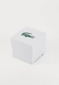 Baltas kartoninis dėžutė su tekstūruotu paviršiumi, ant viršaus esančiu žaliu krokodilo logotipu ir šone užrašytu "LACOSTE".