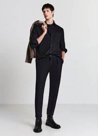 Schwarze Button-Down-Hemd mit einer Tasche, kombiniert mit maßgeschneiderten schwarzen Hosen und schwarzen Stiefeletten. Das Modell hält eine beige Jacke über die Schulter.